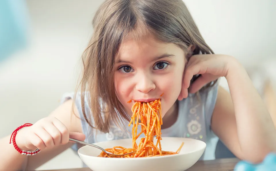 Young girl eating spaghetti with tomato sauce from a white bowl at a table.