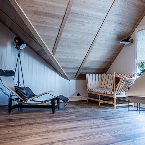 Cozy attic room with slanted wooden ceiling, featuring a black lounge chair, a striped loveseat, and soft lighting.
