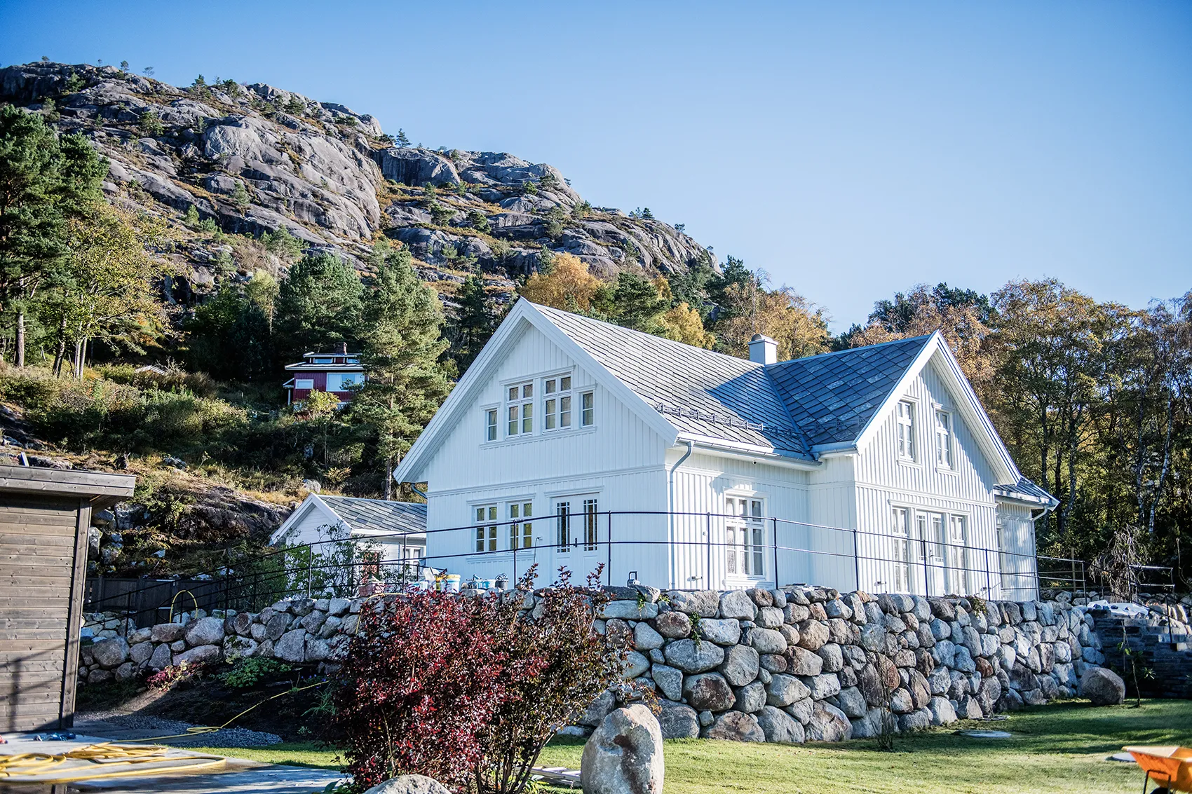 White wooden house with a stone retaining wall, set against rocky hills and trees under a clear blue sky.