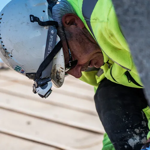 Construction worker wearing a white helmet and neon yellow jacket working closely on a wooden surface.