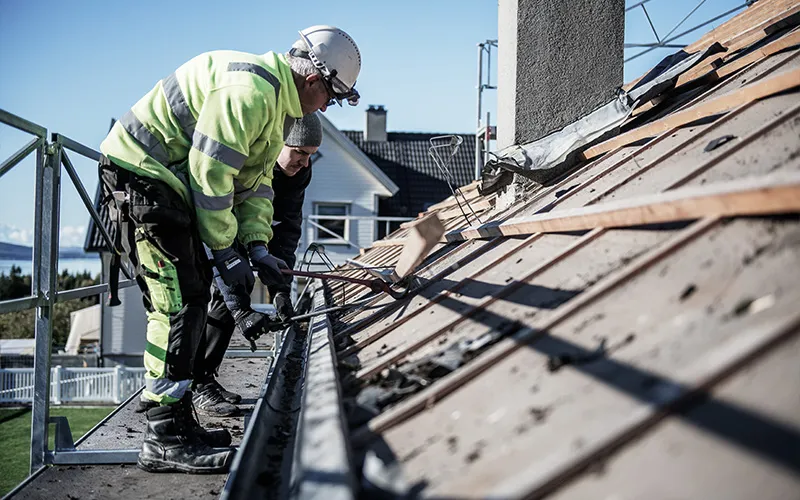 Two construction workers repairing a roof on a sunny day, one wearing a white helmet and neon green jacket.