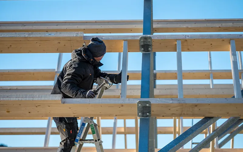 Construction worker in black winter clothing standing on a ladder, installing or inspecting wooden and metal framework under clear blue sky.