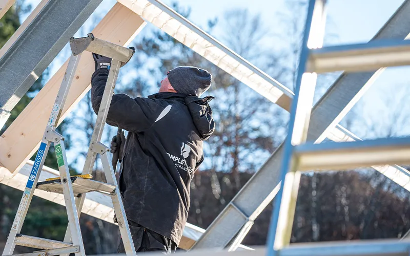 Worker in a black jacket and beanie assembling a wooden frame on a metal structure outdoors.