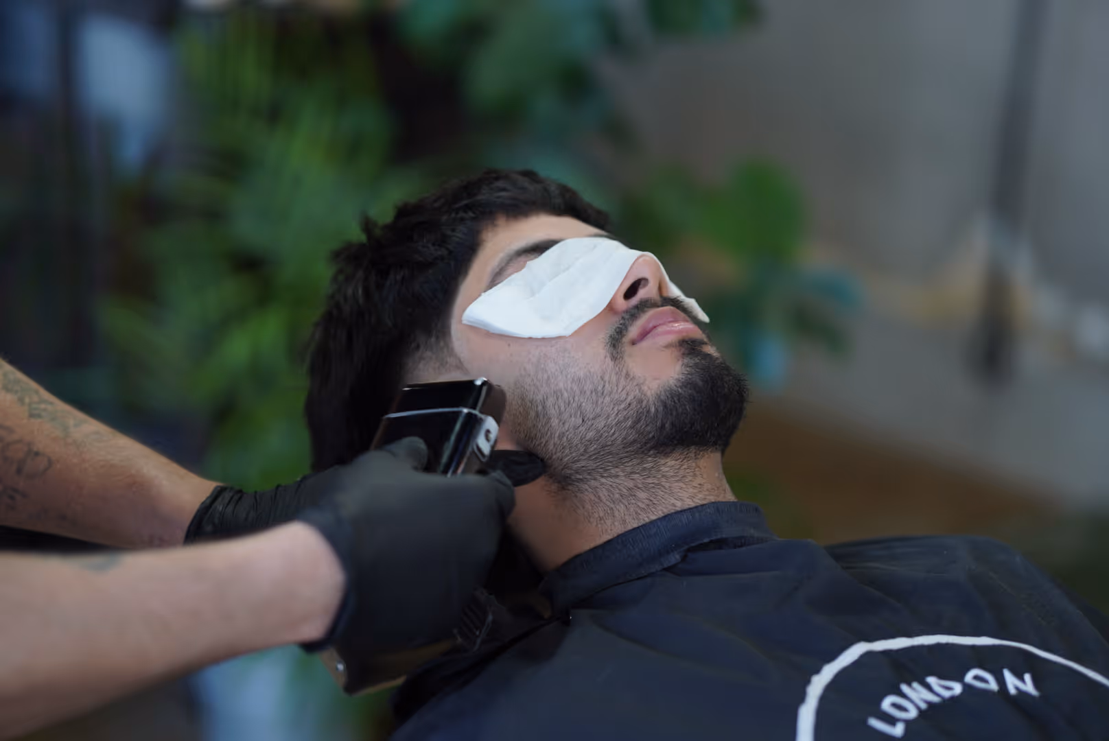 Man with a beard receiving a haircut with electric clippers, eyes covered with a white cloth.