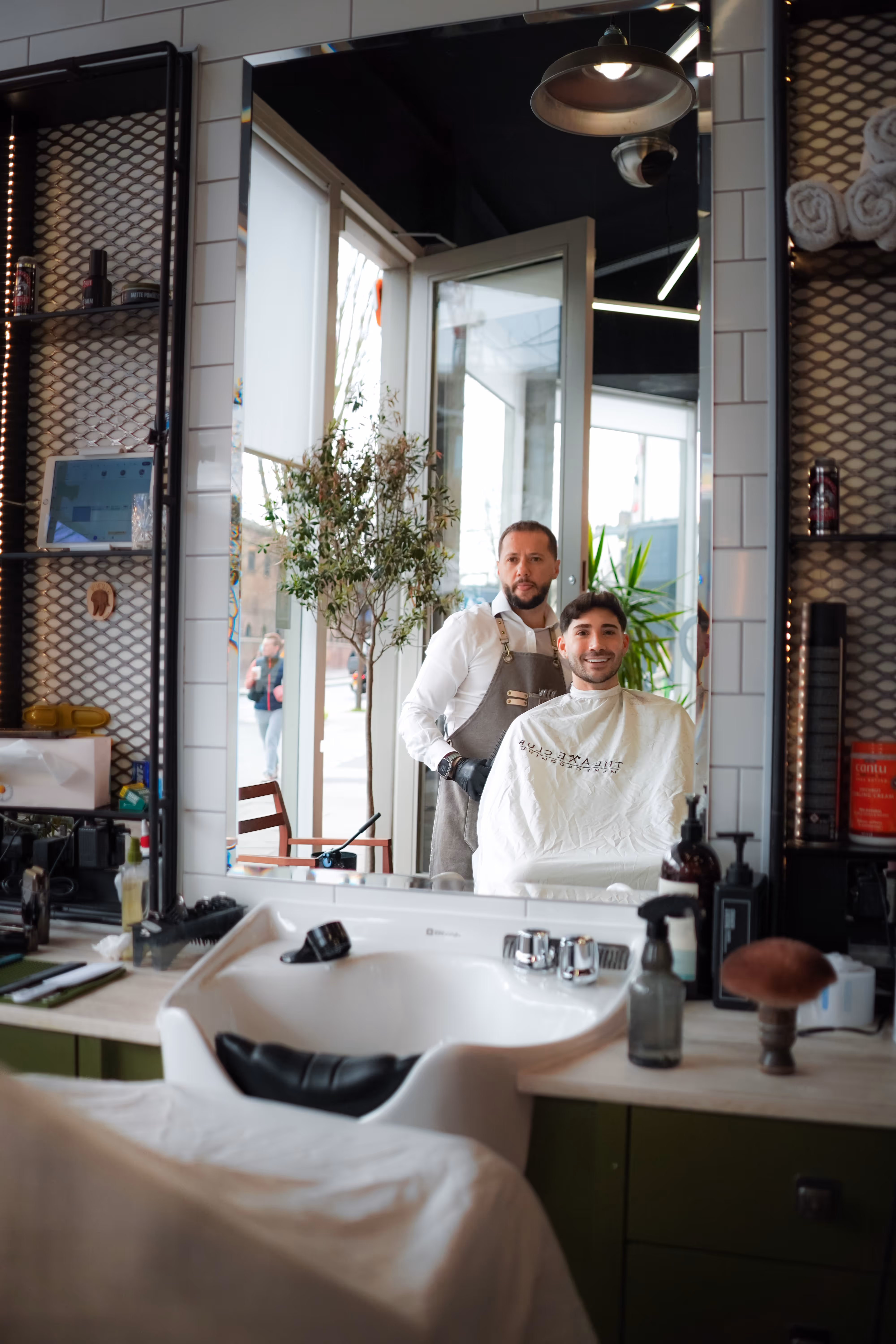 Barber standing behind a smiling man wearing a white cape in front of a salon mirror.