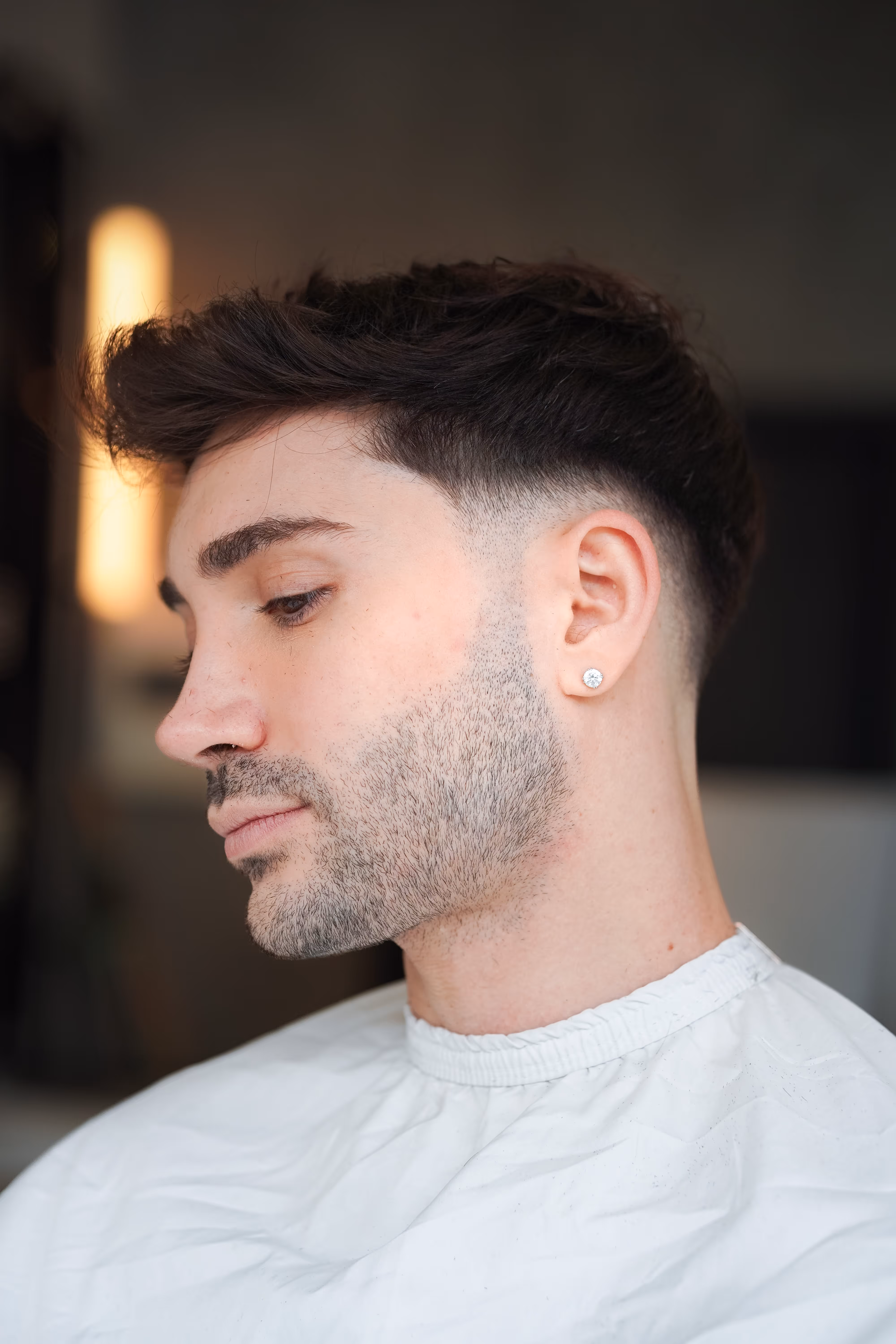 Side profile of a man with dark hair, a trimmed beard, and a diamond stud earring wearing a white barber cape.