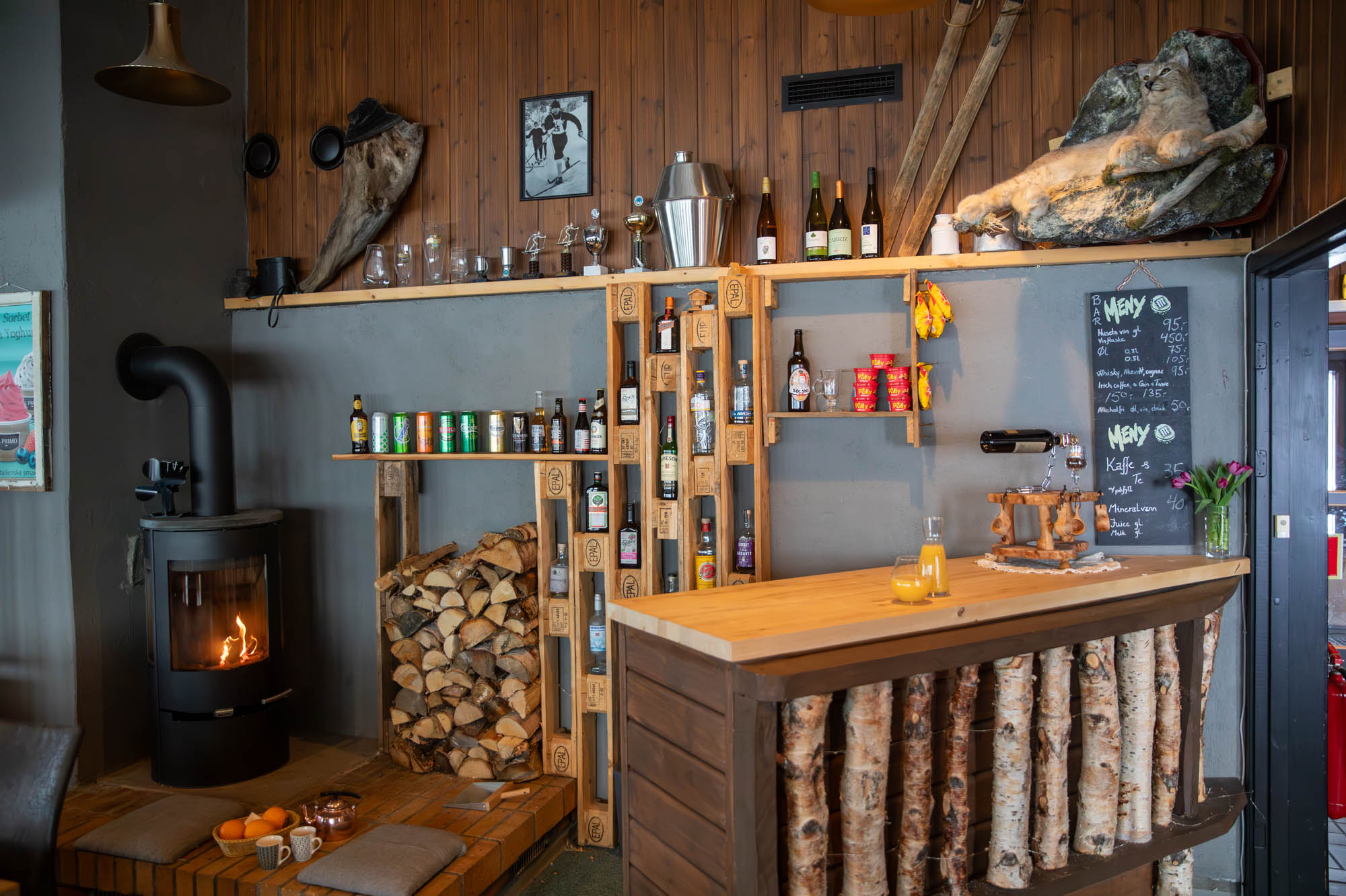 Koselig interiør med peis, vedstabel, bar laget av trestokker og hyller med flasker og drikkevarer, samt menyer på veggen.