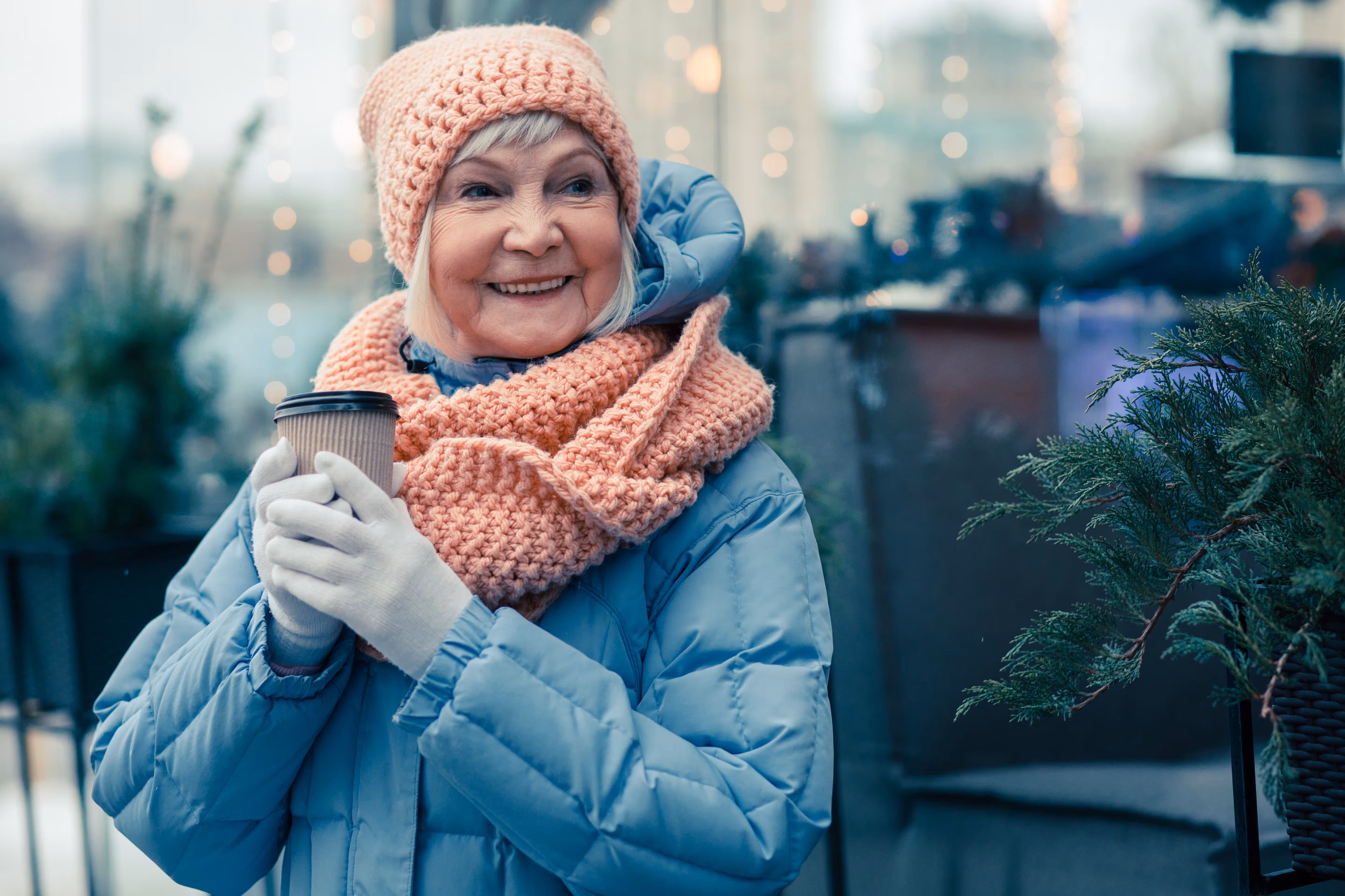 Elderly woman in a blue jacket, pink knit hat and scarf holding a cup of coffee outdoors with festive lights in the background.