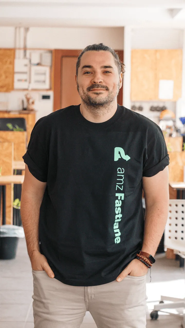 Man with a beard wearing a black amzFastlane t-shirt and beige pants standing in a modern office with hands in pockets.