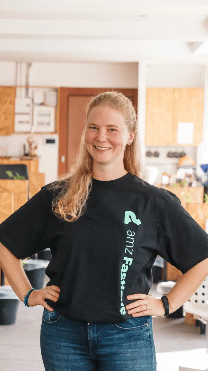 Smiling woman with long blonde hair wearing a black t-shirt with the text 'amz Fastlane', standing with hands on hips in a bright room.