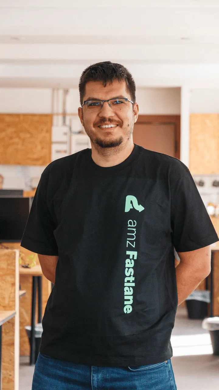 Smiling man wearing glasses and a black t-shirt with amz Fastlane logo standing indoors.