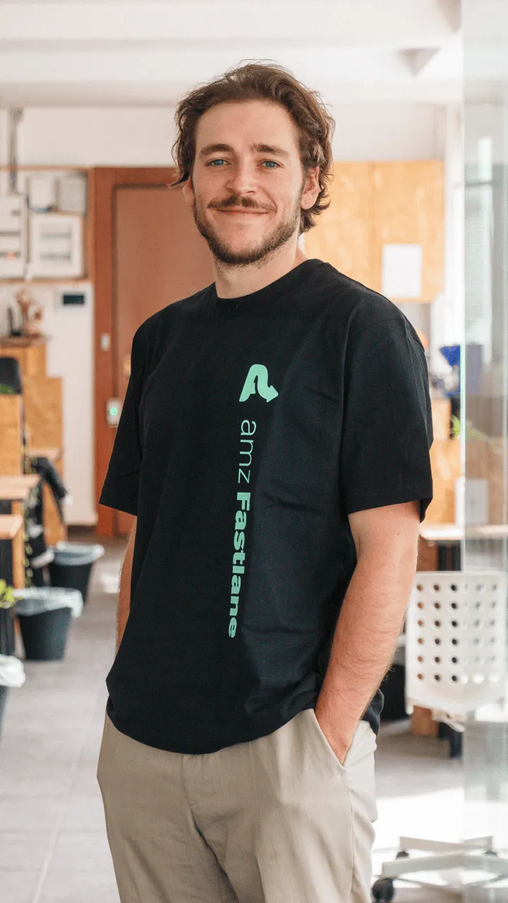 Young man with brown hair and beard wearing a black amz Fastlane t-shirt and beige pants, standing indoors with hands in pockets and smiling.