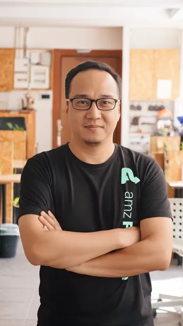 Man with glasses and short black hair wearing a black t-shirt with crossed arms standing in a workspace.