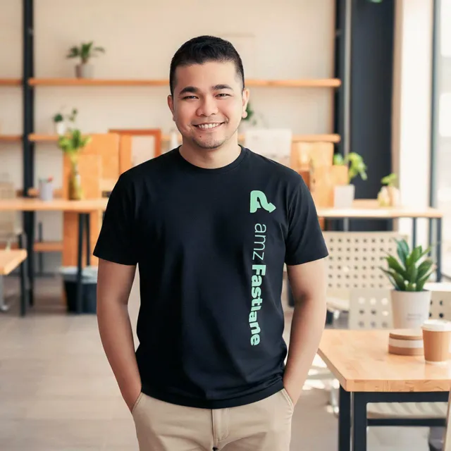 Young man smiling, wearing a black amz Fastlane t-shirt, standing in a modern café with wooden tables and plants.