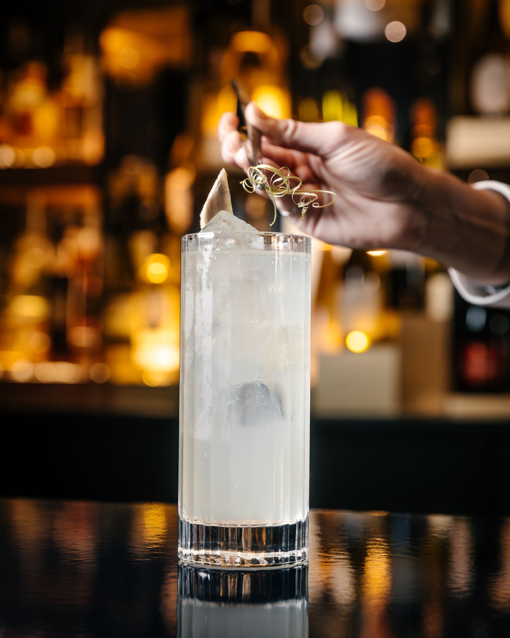 Une main décorant un cocktail clair dans un verre haut avec glace et garniture au bar flou en arrière-plan.