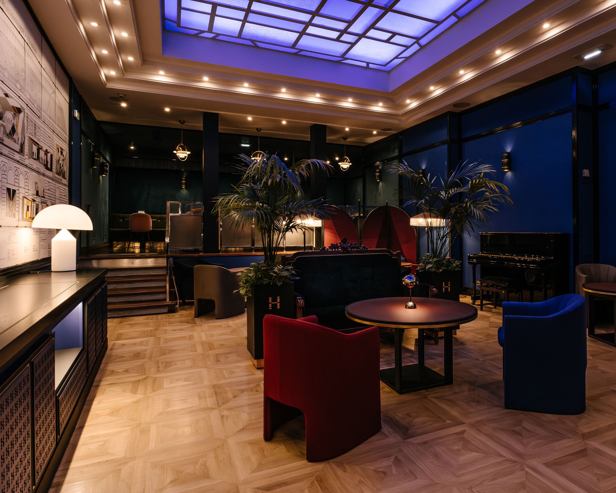 Salon moderne avec sol en parquet clair, fauteuils rouges et bleus autour d'une table ronde, plantes et piano noir dans un éclairage tamisé bleu.