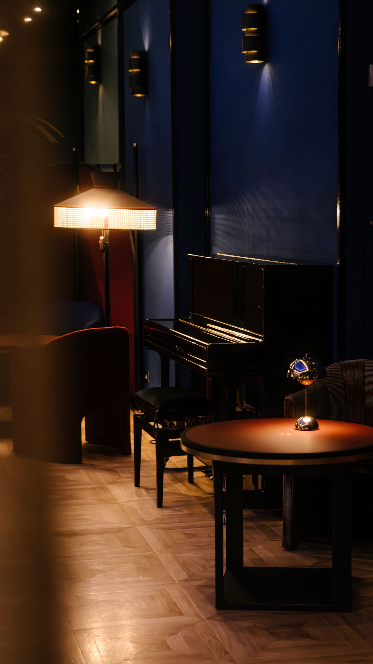 Salon élégant avec un piano noir, une lampe sur pied à lumière tamisée, une table ronde et des fauteuils, murs bleu foncé.