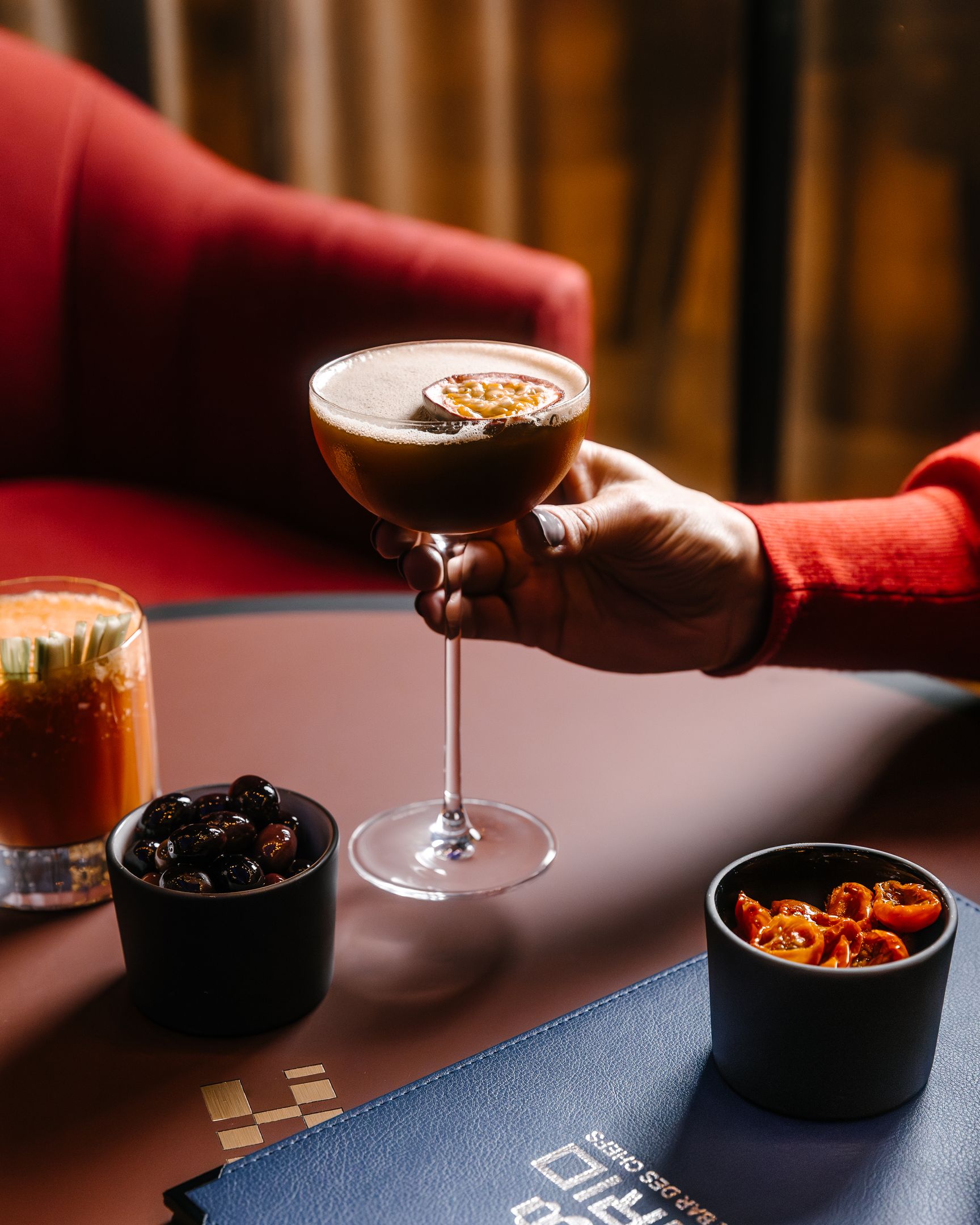 Main tenant un cocktail marron dans un verre à pied avec une demi-passion en garniture, sur une table avec un verre de boisson orange, un bol d'olives noires, un bol de tomates cerises séchées et un menu bleu.