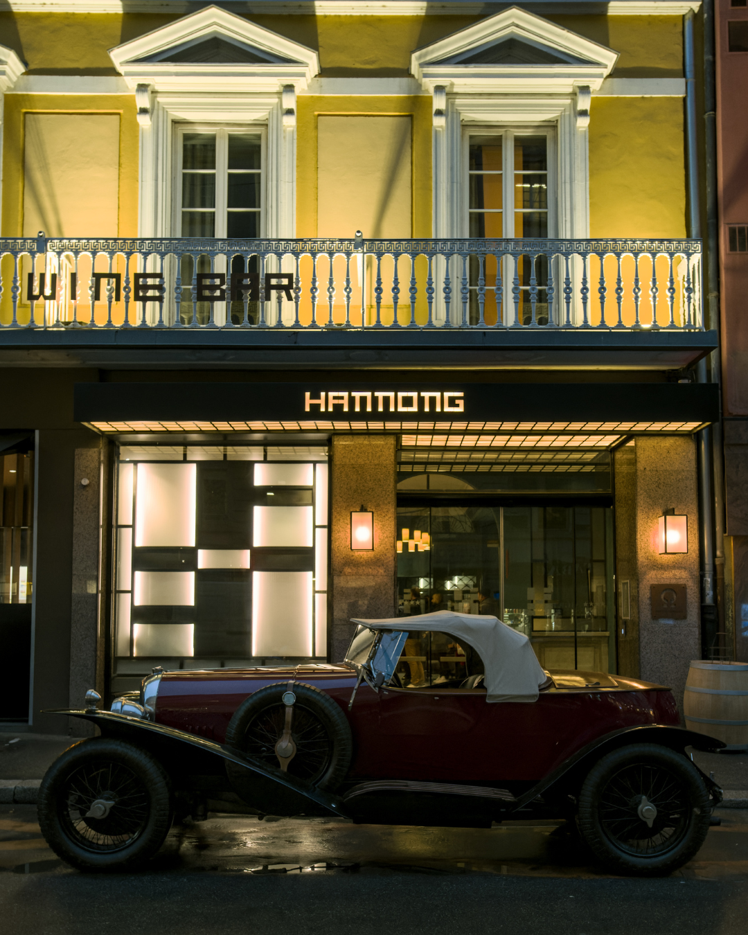 Voiture classique rouge avec toit beige garée devant l'entrée illuminée de l'hôtel Hannong de nuit.