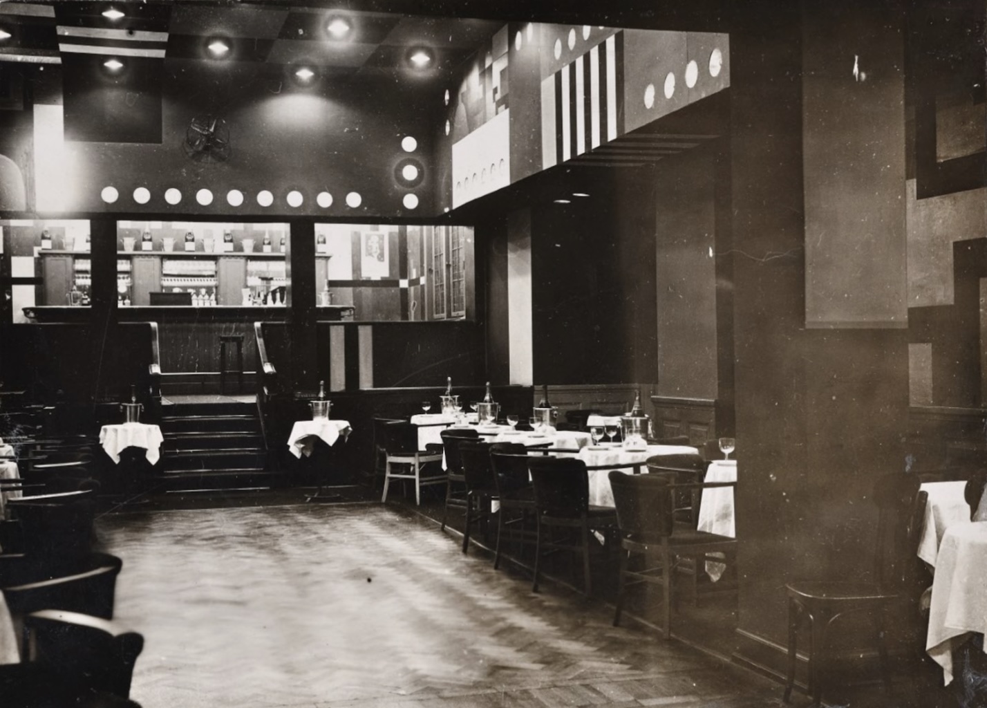 Intérieur d'un cabaret art déco avec tables nappées, chaises, et un bar en arrière-plan.