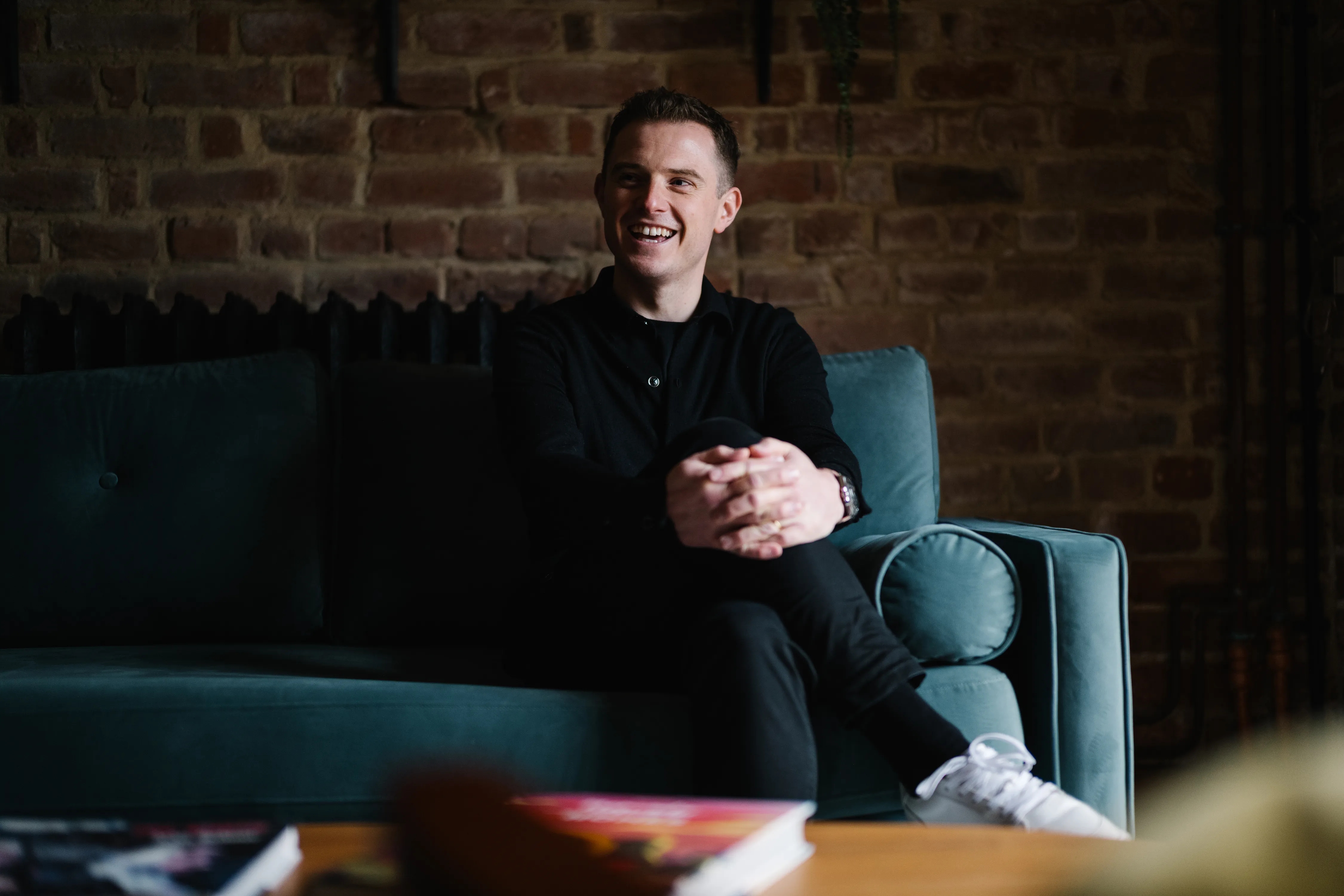 Smiling man in black clothing sitting cross-legged on a teal sofa in front of a brick wall.