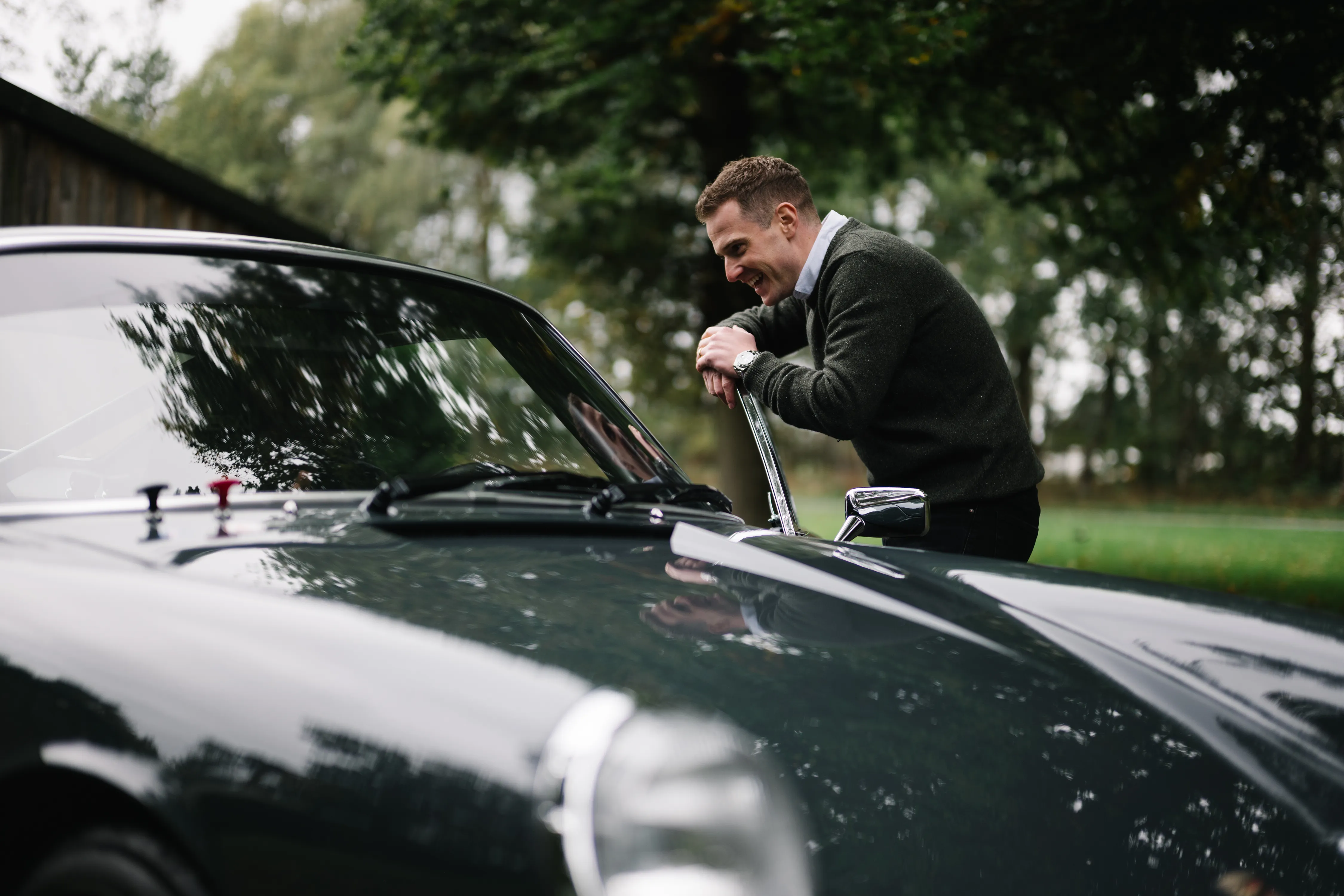 Man leaning on a dark green vintage car outdoors, smiling and looking inside the windshield.