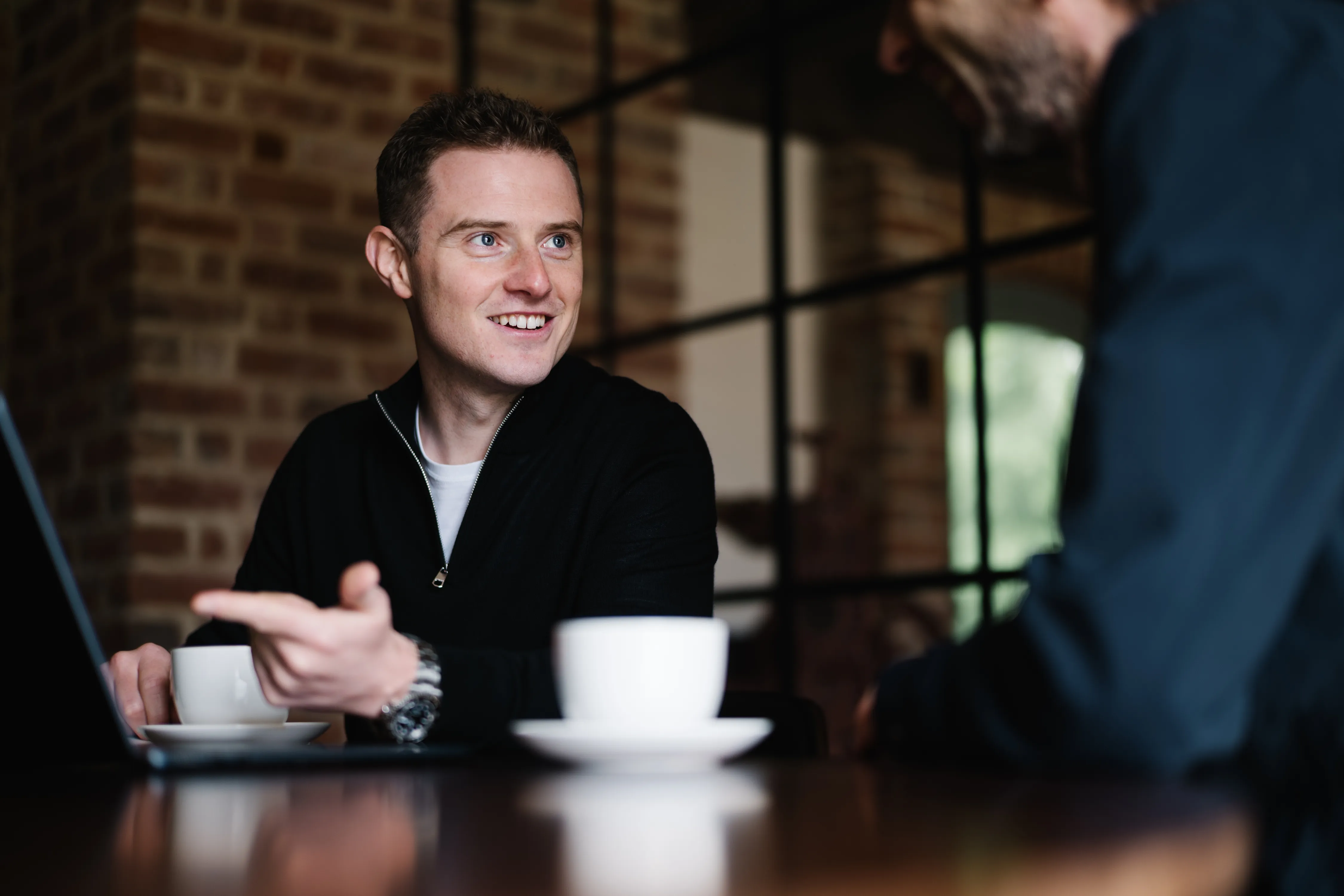 Smiling man in a black zip-up sweater gestures while talking to another person across a table in a cafe.