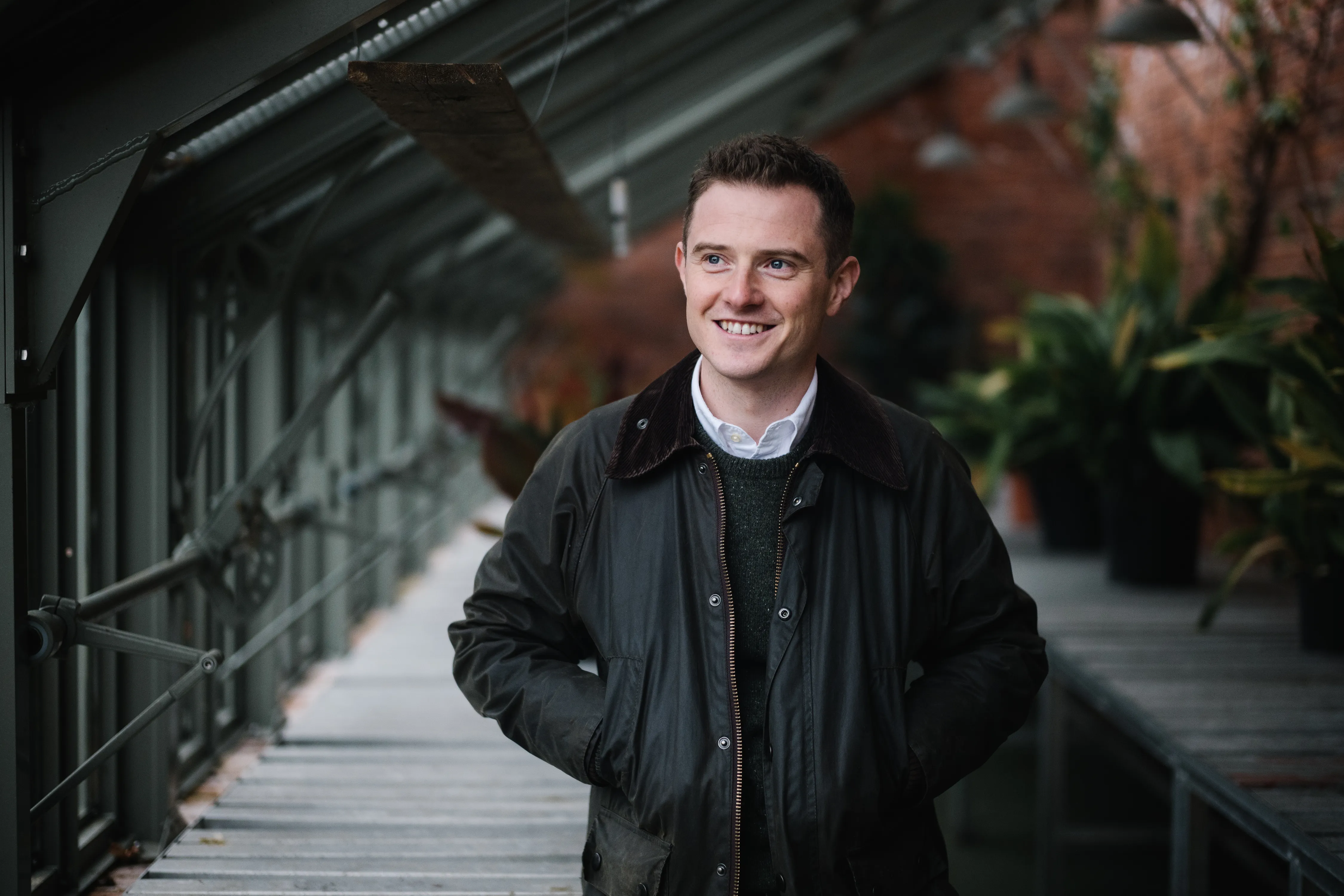 Smiling man with short hair wearing a dark jacket standing in a covered walkway with plants and brick wall in the background.