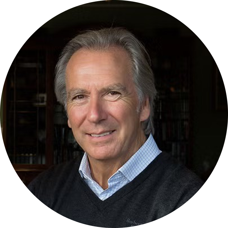 Smiling older man with grey hair wearing a blue checkered shirt under a dark sweater.