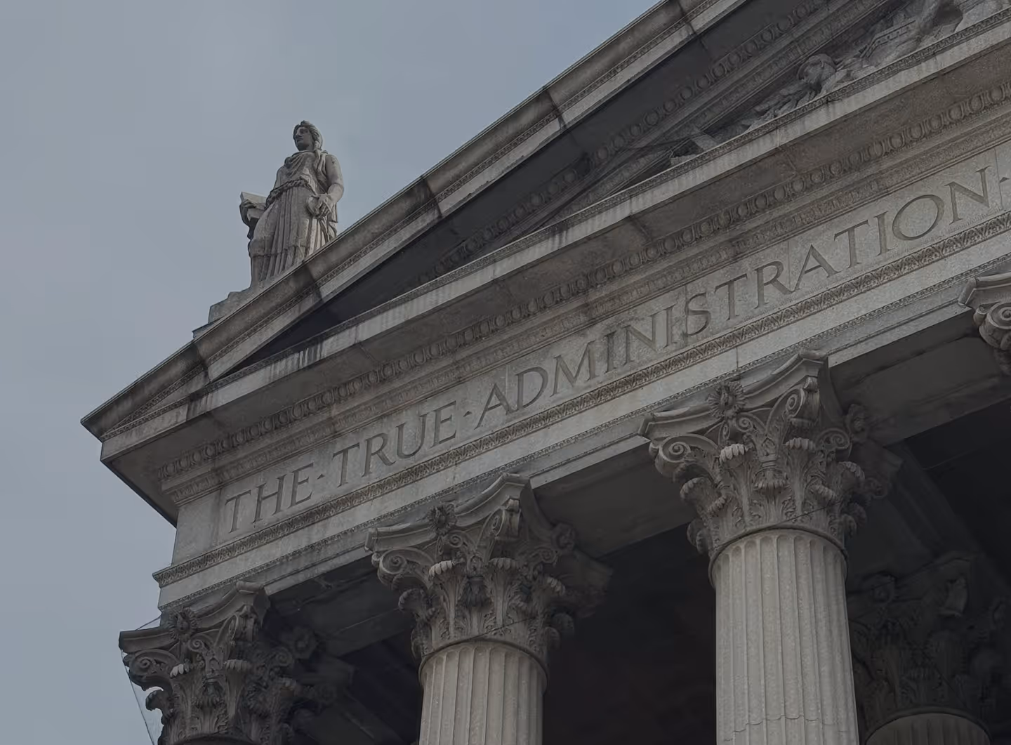 Close-up of a classical building facade with Corinthian columns and a statue on top under a cloudy sky.