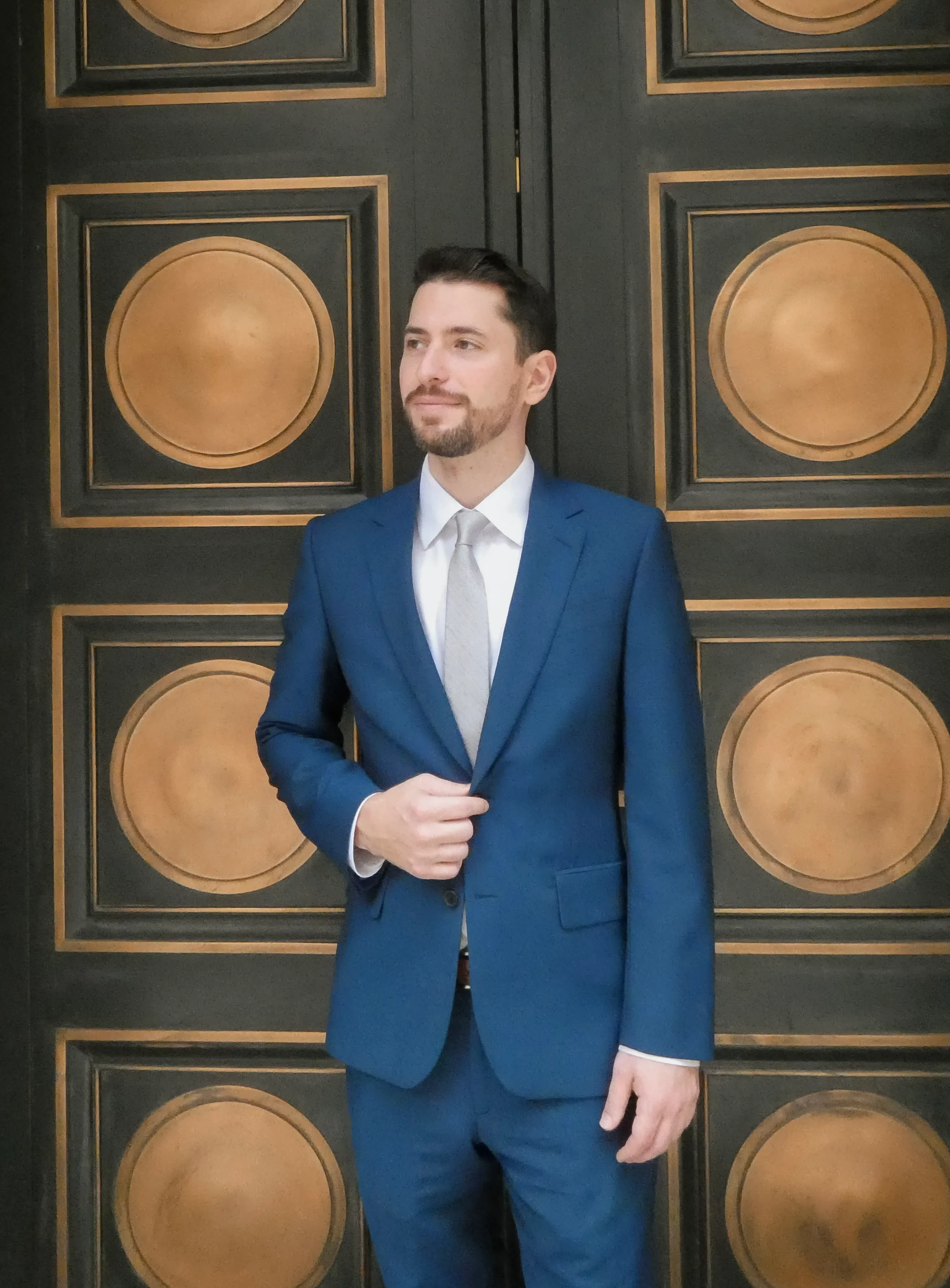 Man in a blue suit with a beige tie standing in front of a black door with gold circular designs.