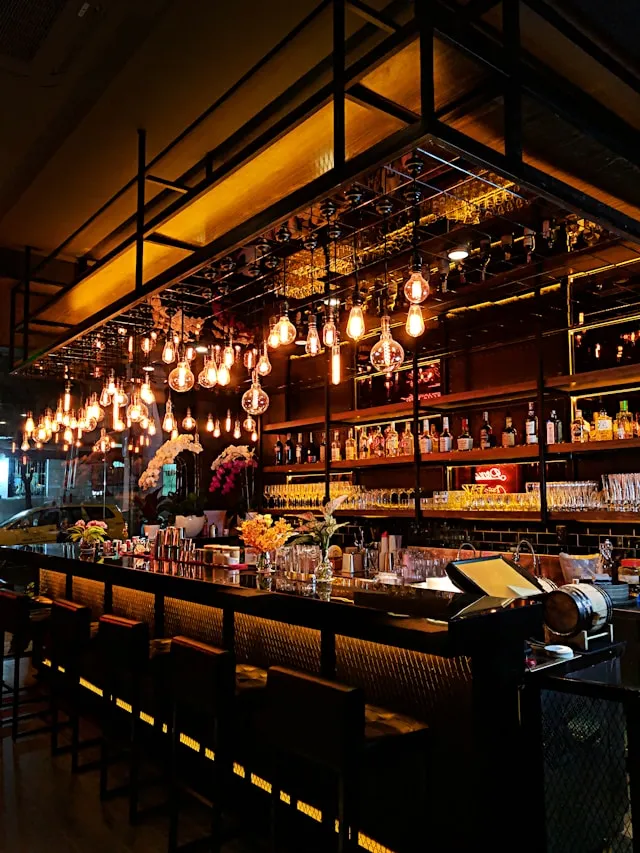 Cozy bar interior with hanging Edison bulbs, shelves of liquor bottles, and a row of stools along the counter.