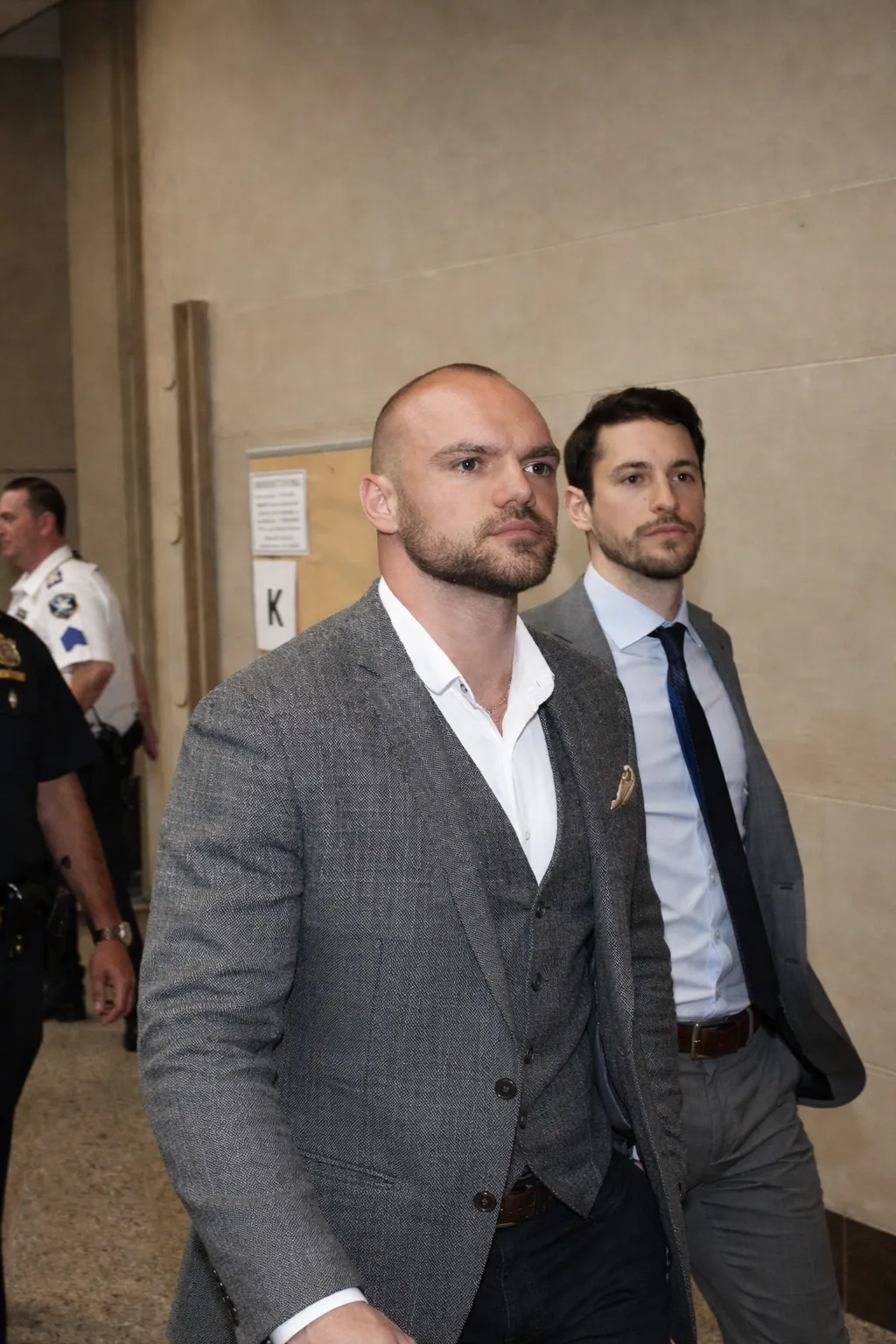 Two men in gray suits walking indoors with police officers visible in the background.