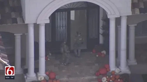 Two people wearing tactical clothing walking through a covered porch area with white columns and pumpkin decorations.