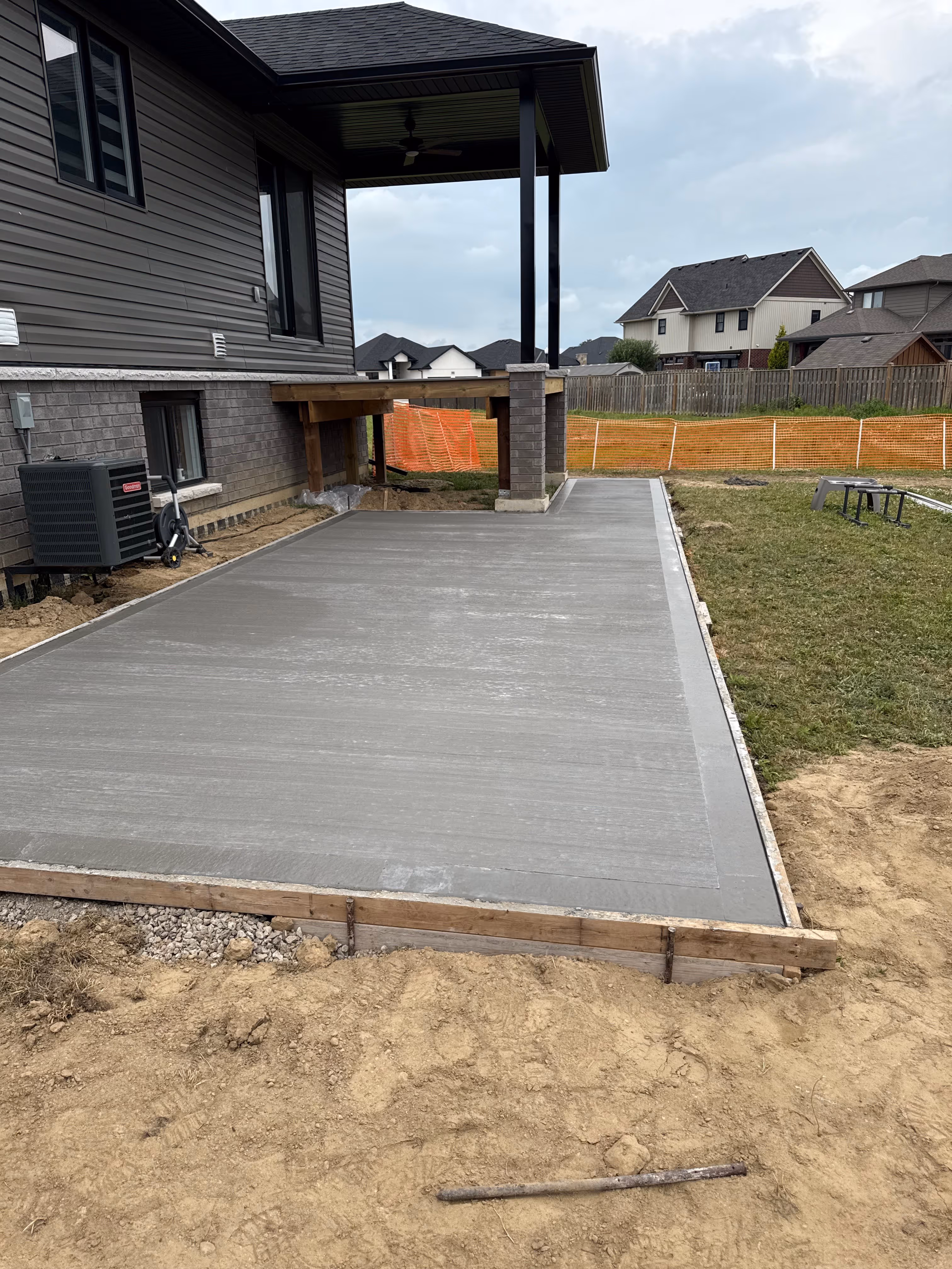 Newly poured concrete patio next to a house with dark siding and brick, bordered by dirt and grass in a residential neighborhood.