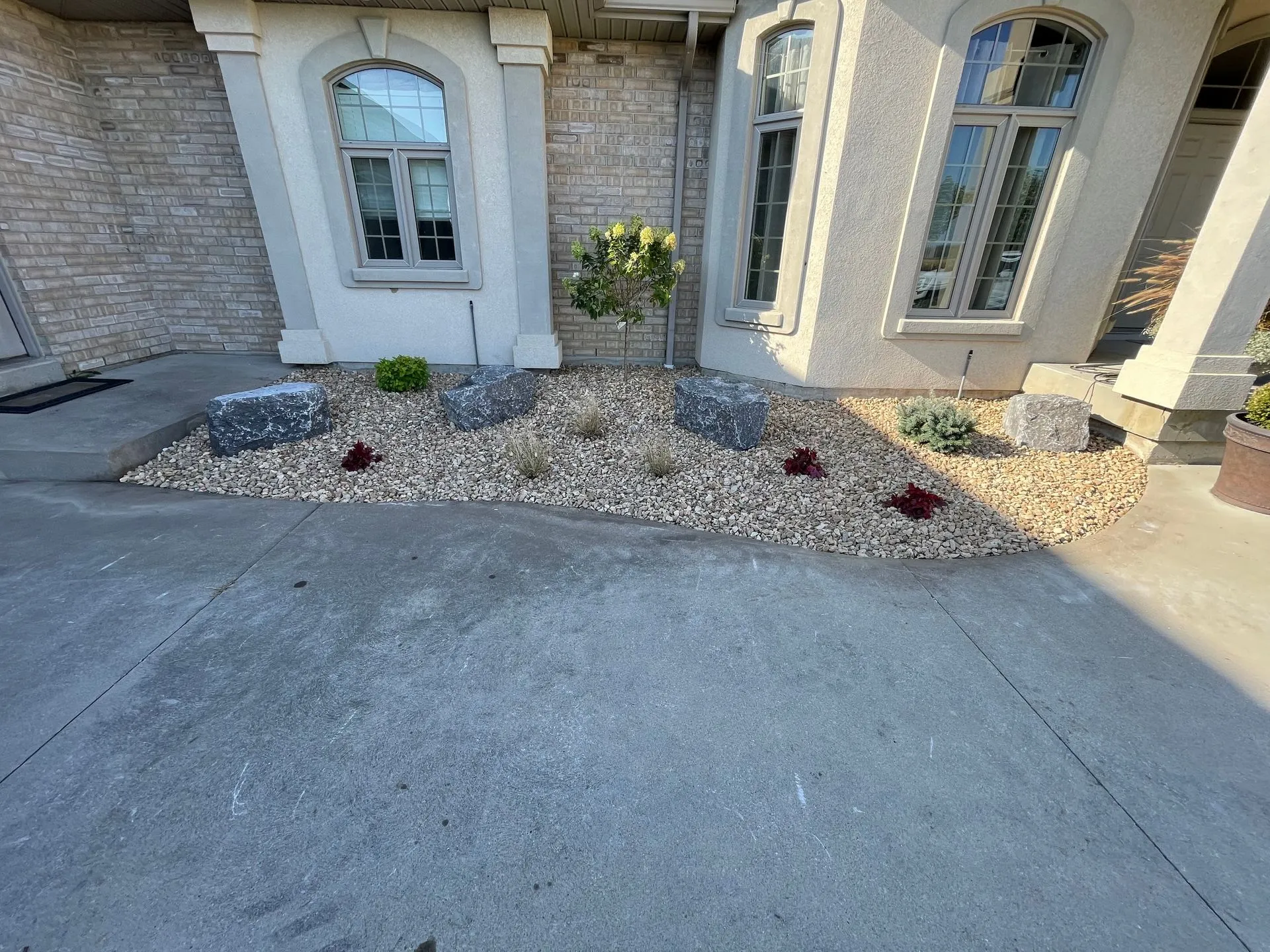 Front yard rock garden with large stones, small shrubs, and a young tree in front of a house with beige brick and stucco exterior.