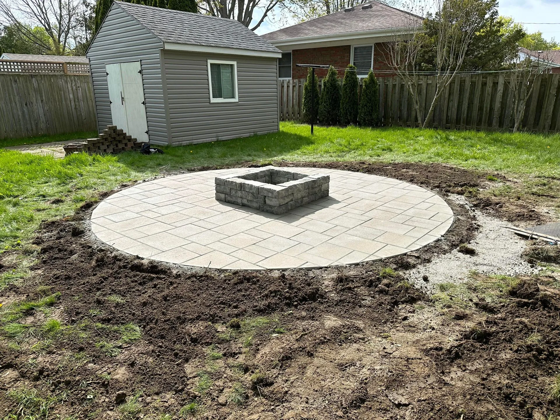 A circular stone patio in a backyard with a square stone fire pit in the center, surrounded by soil and grass, with a shed and wooden fence in the background.