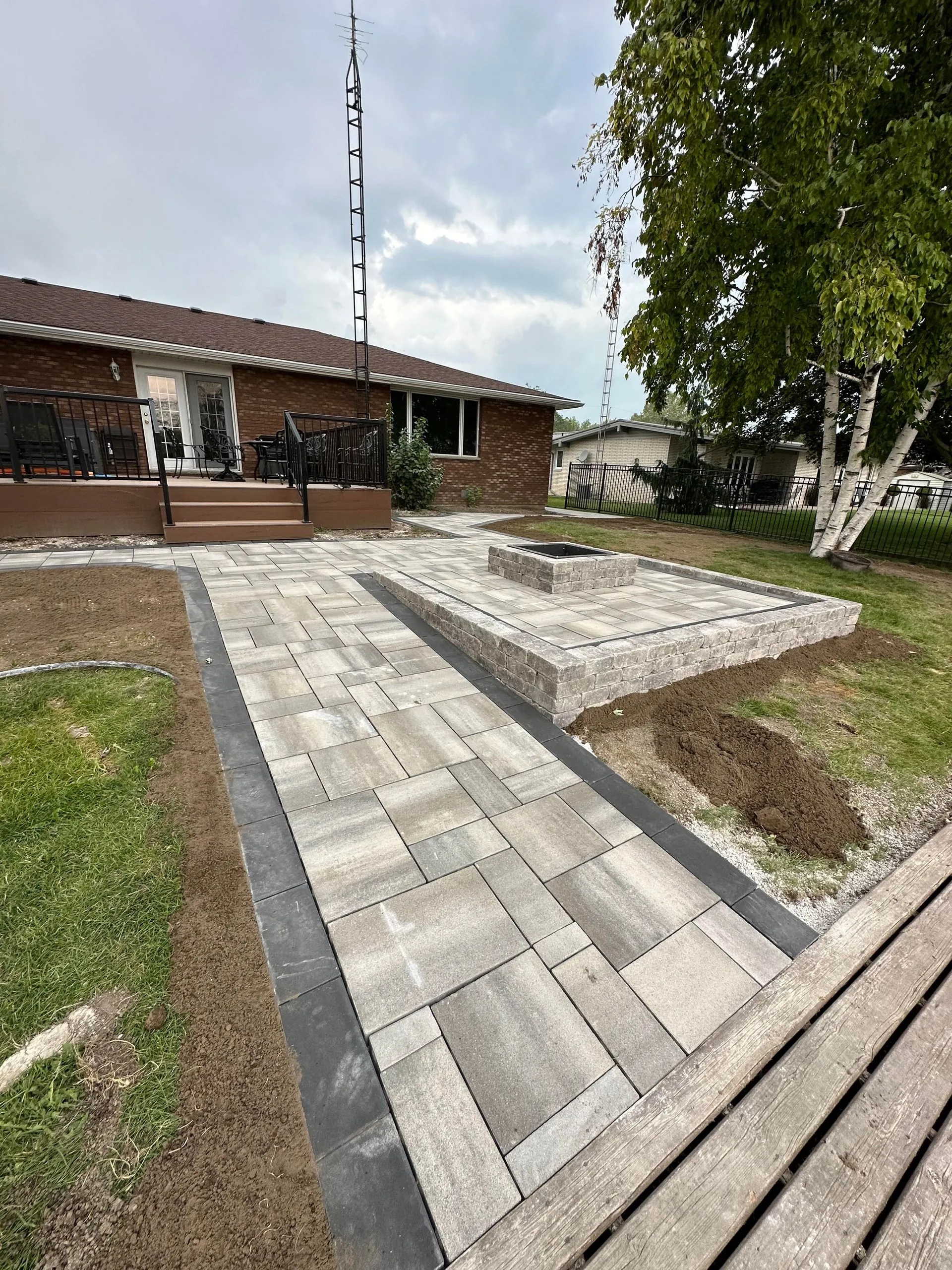 Backyard patio with gray stone pavers, raised fire pit, and a brown brick house with a small deck and black railing.