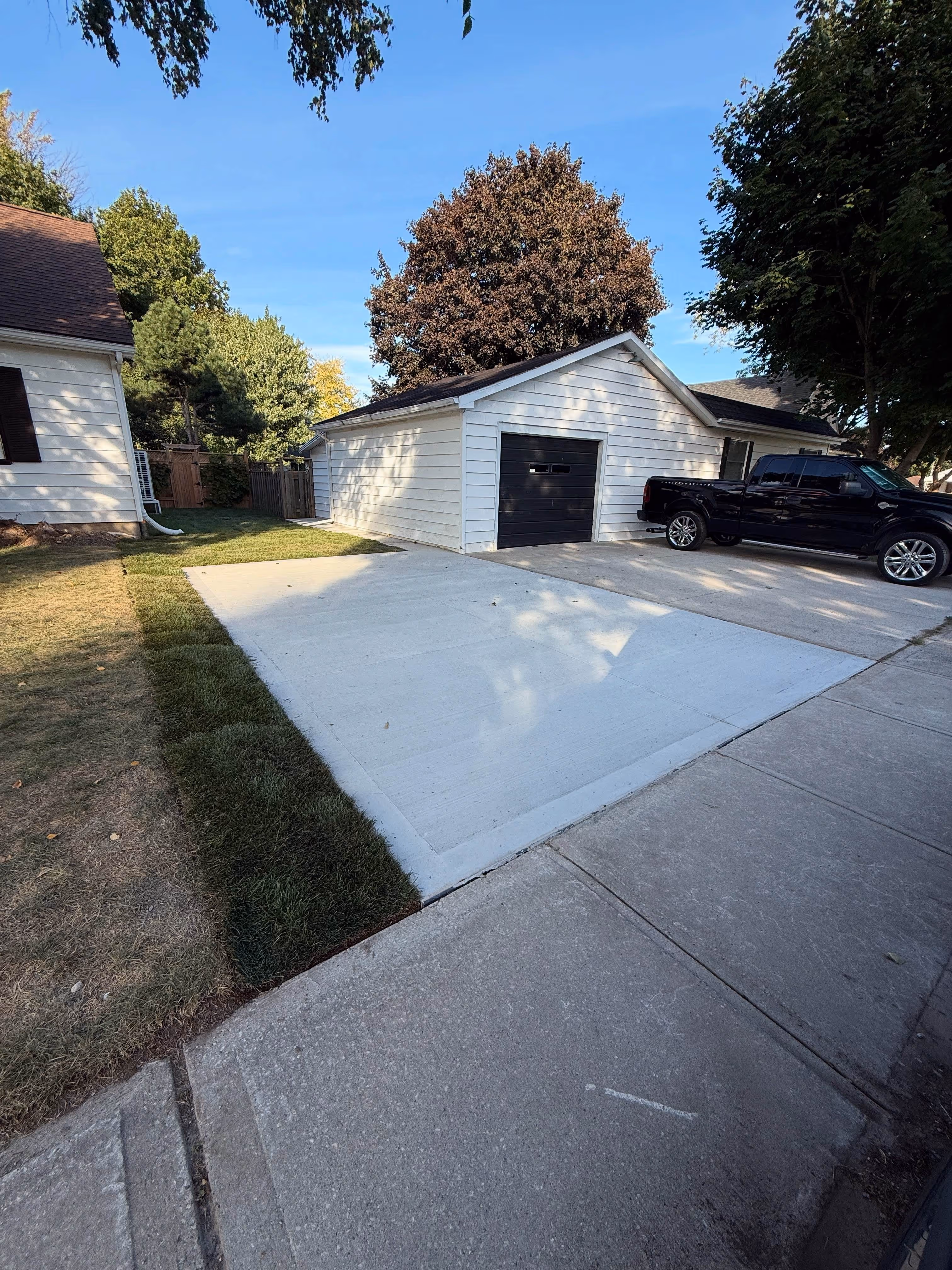Newly poured concrete driveway next to a white garage with black door and a black pickup truck parked beside it.
