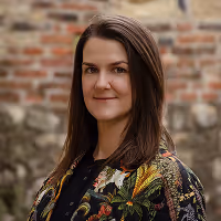 Žena s dlouhými tmavými vlasy a vzorovaným kabátem stojící před cihelnou zdí.