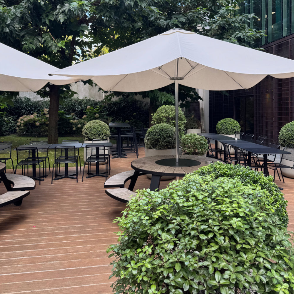 Outdoor seating area with round and rectangular tables, large white umbrellas, and green shrubbery on wooden decking.