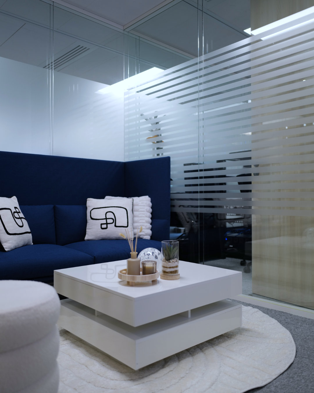 Modern office lounge area with a blue sofa, white abstract-patterned pillows, white round coffee table with decorative items, and frosted glass partition.