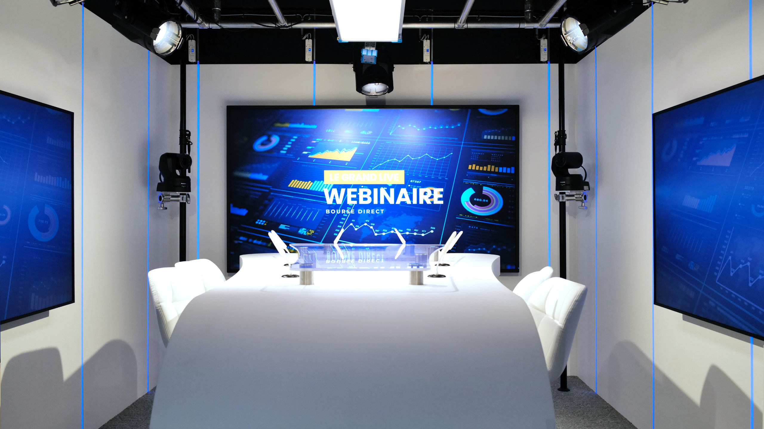 Modern webinar studio with a curved white table, white chairs, microphones, and three large screens displaying digital data and the text 'Le Grand Live Webinaire Bourse Direct'.