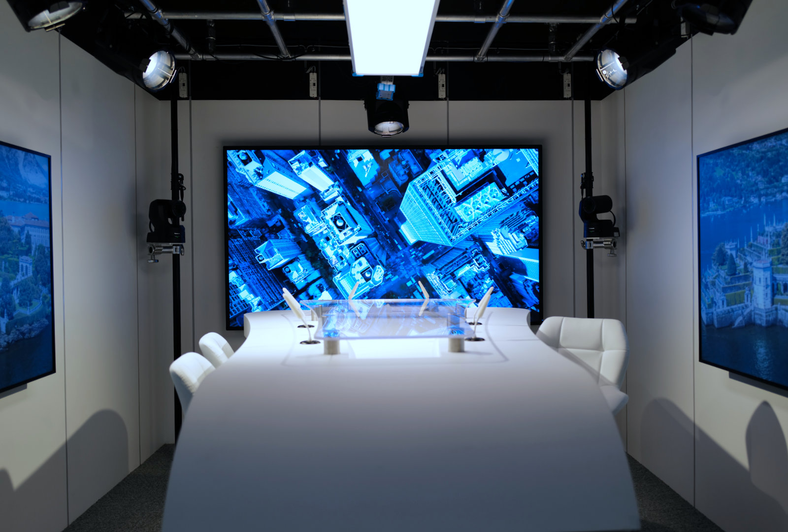 Modern conference room with curved white table, white chairs, and large digital screens showing aerial cityscape and waterfront images.