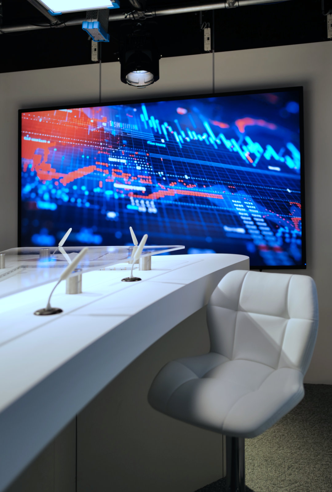 Modern white chair next to a curved white desk with microphones, in front of a large screen displaying colorful digital data graphs.