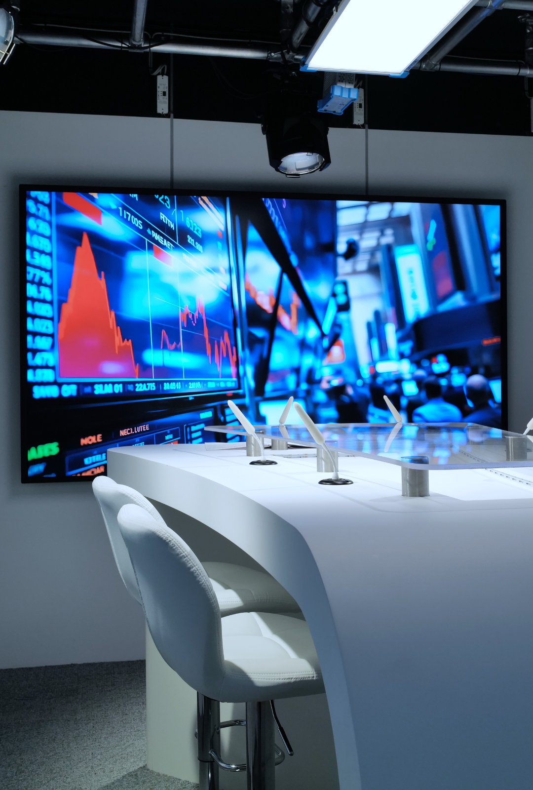 Modern white curved desk with stools in front of a large screen displaying stock market graphs and trading data in a studio setting.