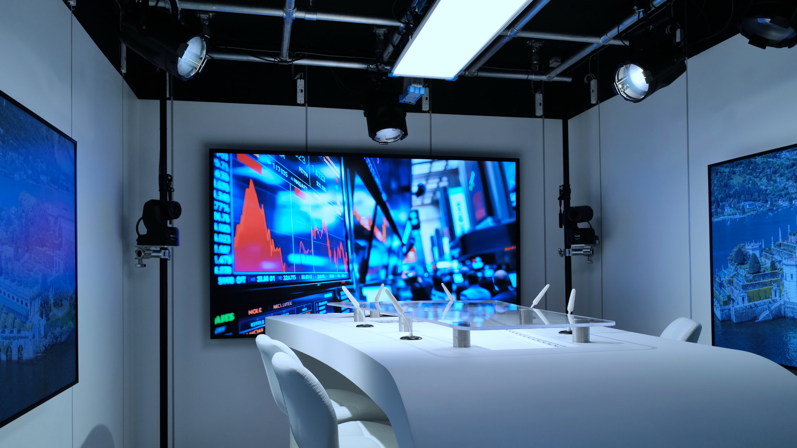Modern studio setup with white chairs around a curved white table and large screens displaying a stock market graph and a scenic aerial view.
