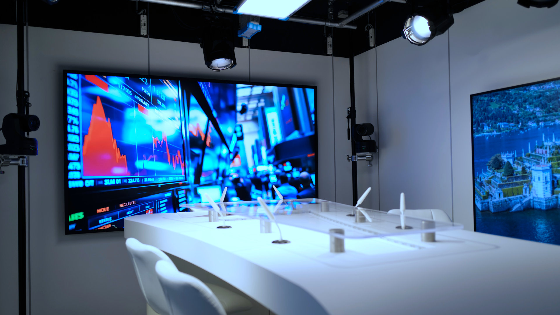 Modern conference room with white table and chairs, large screen showing blue and red financial charts, and another screen displaying an aerial view of a castle by a lake.