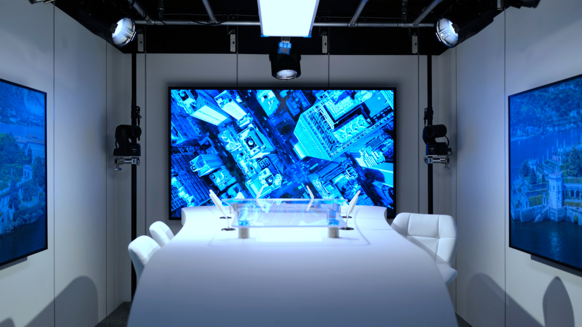 Modern conference room with white curved table and chairs, featuring a large screen showing a blue aerial cityscape view, flanked by two smaller screens displaying scenic landscapes.