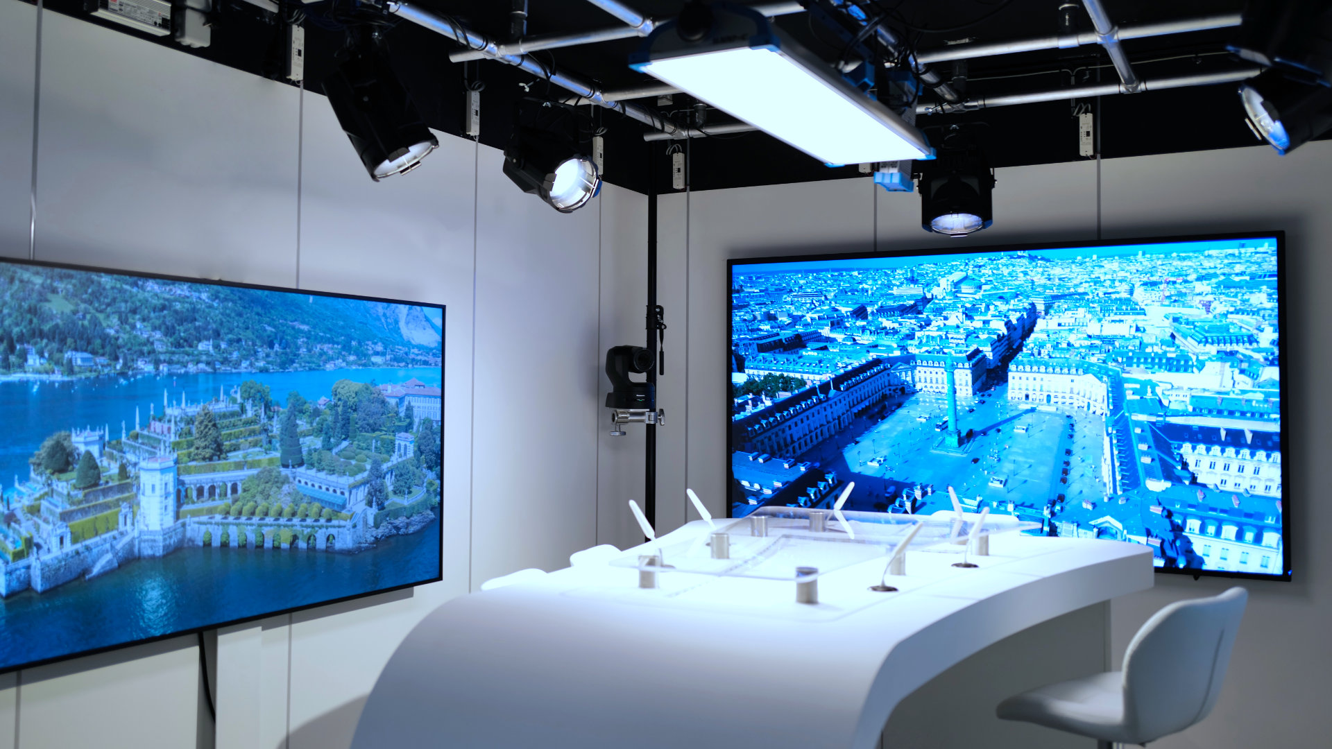 Modern studio with two large screens showing aerial city views and a curved white desk with microphones and a chair.